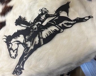 May include: Black metal silhouette wall art depicting a cowboy riding a bucking bronco. The cowboy is wearing a hat and has a lasso in his hand.