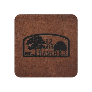 May include: Brown leather coaster with a black silhouette of two trees, a cow, and a fence. The text "1203 RANCH" is also included in the design.