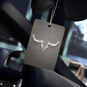 May include: Rectangular car air freshener with a gray background and a white bull skull design. The air freshener hangs from a white string. The image is set inside a car.