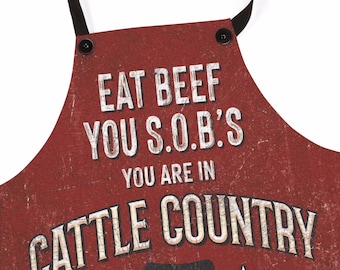 Op de afbeelding: Rode en witte schort met de tekst "Eat Beef You S.O.B.'s You Are In Cattle Country" en een zwart silhouet van een koe met twee witte sterren.
