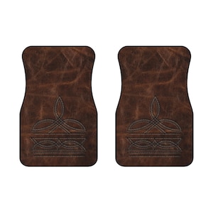 May include: Two brown car floor mats with a leather-like texture. Each mat features a symmetrical, dark brown embroidered design. The design includes a stylized crown-like shape at the bottom and a looping pattern at the top.