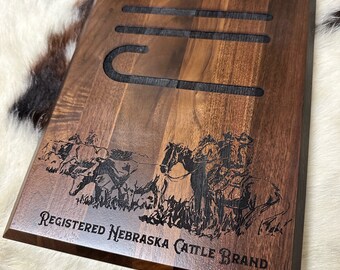 May include: A wooden plaque with a black engraved design of a cowboy riding a horse and cattle. The plaque reads "Registered Nebraska Cattle Brand".