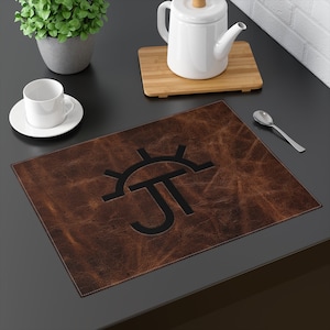 Op de afbeelding: Een rechthoekige bruine leren placemat met een zwart paraplu-achtig logo. De placemat heeft een getextureerd oppervlak en witte stiksels langs de randen. Een witte kop en schotel, een theepot en een lepel zijn ook zichtbaar.