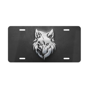 May include: A black license plate featuring a silver wolf head design. The wolf's head is centered and detailed, with a textured background. The plate has four mounting holes, and the overall design is sleek and modern.