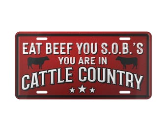 May include: Red metal sign with white text that reads "Eat Beef You S.O.B.'s You Are In Cattle Country" with two black silhouettes of cows on either side of the text. There are three white stars below the text.