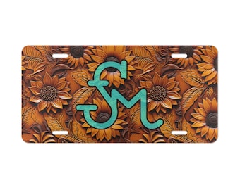 May include: A brown leather license plate with a turquoise blue monogram "SM" in the center. The background features a repeating pattern of sunflowers.