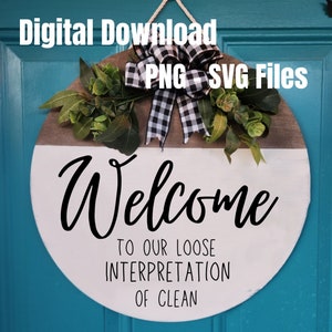 May include: A round wooden welcome sign with a white and brown color scheme. The sign features the word "Welcome" in large, black cursive letters, along with the phrase "TO OUR LOOSE INTERPRETATION OF CLEAN." It is decorated with greenery and a black and white checkered bow.