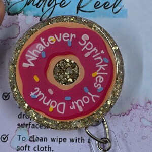 May include: A badge reel with a pink donut design. The donut has sprinkles and the words "Whatever Sprinkles Your Donut" in a circular pattern. The reel has a glittery gold border and a clear plastic clip.