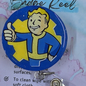 May include: A blue badge reel featuring a cartoon character giving a thumbs up. The character is wearing a blue jumpsuit and has blonde hair. The badge reel has a clear plastic strap and a metal clip.