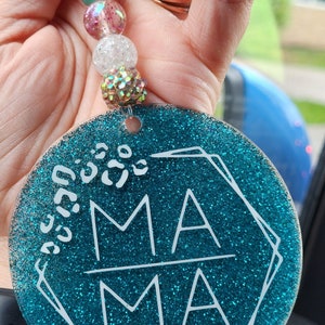 May include: A teal glitter acrylic car charm with the word "MAMA" in white, accented with a geometric design and leopard print spots. The charm is attached to a beaded strand with various colorful beads.