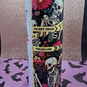 May include: A stainless steel tumbler with a black floral and skeleton design. The tumbler features red flowers, a "Police Line" and "Do Not Cross" warning tape design. The tumbler is cylindrical and has a white stripe on the side.