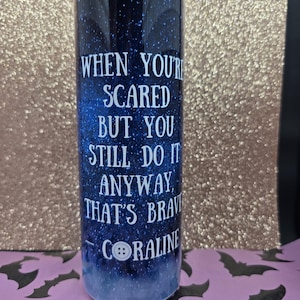 Pode incluir: Um copo cil&iacute;ndrico com um fundo azul escuro e estrelado. O texto branco diz: "QUANDO VOC&Ecirc; EST&Aacute; COM MEDO, MAS AINDA ASSIM FAZ, ISSO &Eacute; CORAJOSO CORALINE". Uma imagem de bot&atilde;o branco est&aacute; abaixo do texto. O copo tem cerca de 20 cm de altura.