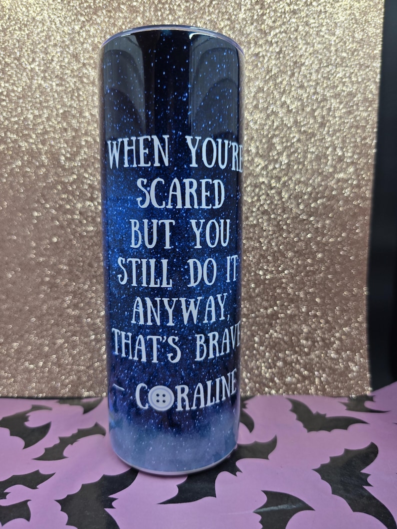 Pode incluir: Um copo cil&iacute;ndrico com um fundo azul escuro e estrelado. O texto branco diz: "QUANDO VOC&Ecirc; EST&Aacute; COM MEDO, MAS AINDA ASSIM FAZ, ISSO &Eacute; CORAJOSO CORALINE". Uma imagem de bot&atilde;o branco est&aacute; abaixo do texto. O copo tem cerca de 20 cm de altura.