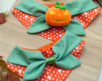 May include: Two Halloween-themed dog bandanas. Each bandana features a green bow on an orange fabric with white polka dots and ghost designs. One bandana has a plush orange jack-o'-lantern in the center.
