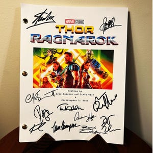 Può includere: Una copertina di sceneggiatura bianca per il film "Thor: Ragnarok" con il titolo in un font retrò colorato. La copertina presenta una foto dei personaggi principali del film. La copertina è firmata da diverse persone, tra cui Stan Lee.