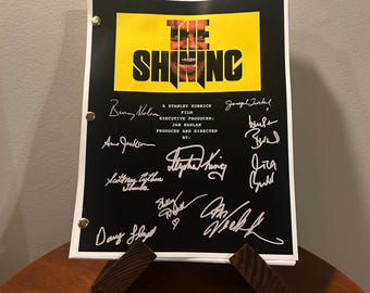 May include: A black and yellow movie poster for the film "The Shining" with the title in bold white letters. The poster features a close-up of Jack Nicholson's face as Jack Torrance, the main character. The poster is signed by several people, including Stephen King, Jack Nicholson, and Shelley Duvall.
