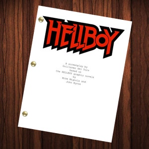May include: A white script with black text on a wooden background. The text reads "HELLBOY" in a bold, stylized font. Below the title, it says "A screenplay by Guillermo del Toro based on the HELLBOY graphic novels by Mike Mignola and John Byrne."