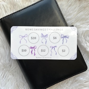 May include: A white card with the text "BOWS SAVINGS CHALLENGE" and illustrations of purple bows. The card has circles with dollar amounts: $20, $6, $10, $12, and $2. The card is on a black leather wallet.
