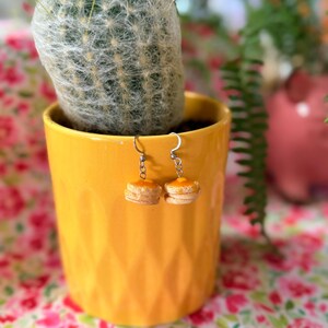 May include: A pair of dangle earrings featuring miniature pancake stacks with a golden syrup topping. The earrings are suspended from silver-toned metal hooks and are displayed hanging in front of a bright yellow ceramic plant pot.