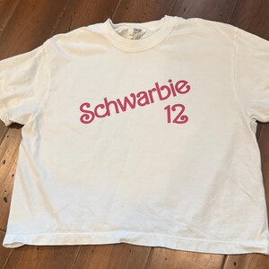 May include: White t-shirt with the text "Schwarbie 12" in a pink, cursive font. The shirt is made of a soft material and has a crew neck. The shirt is laying on a wooden surface.