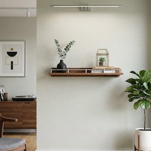 May include: A modern interior scene featuring a wooden shelf with decorative items. A black vase with greenery, a small terrarium, and books are displayed. A framed abstract art piece and a potted plant add to the room's aesthetic. A record player and chair are also visible.