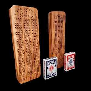 May include: Two wooden cribbage boards with drilled holes and a carved design, accompanied by two decks of Bicycle playing cards. The boards are made of light brown wood and stand upright against a black background.