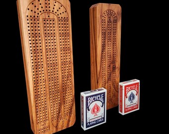 May include: Two wooden cribbage boards with drilled holes and a carved design, accompanied by two decks of Bicycle playing cards. The boards are made of light brown wood and stand upright against a black background.