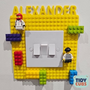 Könnte beinhalten: Eine gelbe Lego-artige Lichtschalterabdeckung mit dem Namen "ALEXANDER" in gelben Buchstaben. Die Abdeckung hat einen weißen Lichtschalter mit drei Kippschaltern. Lego-Figuren und -Blöcke in verschiedenen Farben sind angebracht.
