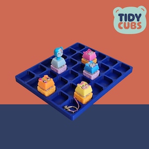 Puede incluir: Un tablero de juego azul con piezas de juguete coloridas. Las piezas son de varios colores, incluyendo naranja, rosa, azul y amarillo, cada una con un personaje de dibujos animados en la parte superior. El tablero tiene el texto "Tidy Cubs" en un logo con forma de oso.