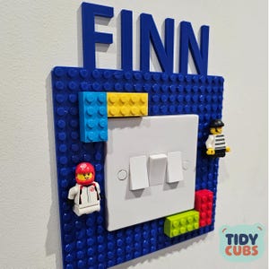 May include: A blue Lego-style light switch cover with the name "FINN" in blue letters above. The cover has colorful Lego bricks and Lego figures attached. The switch plate is white with three switches. The logo "TIDY CUBS" is in the bottom right corner.