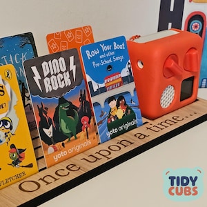 May include: A wooden display stand engraved with "Once upon a time..." holding children's book cards and an orange audio player. The cards display illustrations and text, including "Dino Rock" and "Row Your Boat" with a London bus.