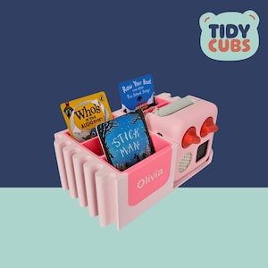 May include: A pink and white Tidy Cubs organiser with compartments holding children's books. The organiser has a handle and red knobs. The books include "Who's in Your Audio Book?", "Row Your Boat", and "Stick Man". The name "Olivia" is printed on the side.