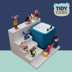 Può includere: Un espositore bianco a più livelli con varie statuette di personaggi, tra cui Spider-Man e Stitch, disposte attorno a un altoparlante blu. L'altoparlante ha una parte superiore bianca con un anello luminoso e due accenti neri. Il testo "Tidy Cubs" è in alto a destra.