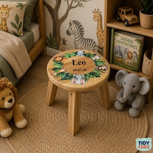 Puede incluir: Un pequeño taburete de madera con una tapa redonda con un diseño de temática selvática. La parte superior tiene el nombre "Leo" y la fecha "18/07/26" impresos. El taburete está rodeado por una alfombra de yute y un juguete de elefante de peluche.