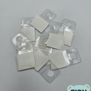May include: A pile of clear plastic adhesive hooks with white adhesive backing. The hooks have a round hole in the center and are designed for hanging items on walls.