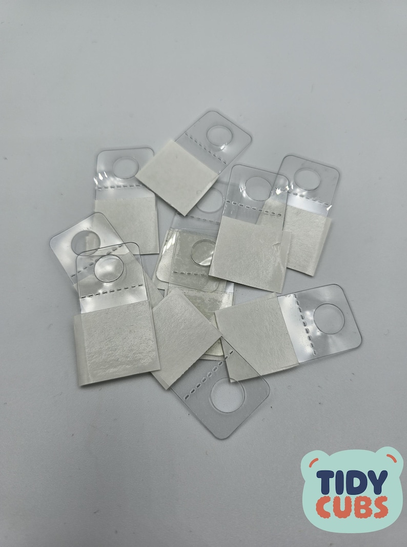 May include: A pile of clear plastic adhesive hooks with white adhesive backing. The hooks have a round hole in the center and are designed for hanging items on walls.