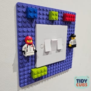 May include: A purple Lego-style wall plate with a white light switch. The plate is decorated with colourful Lego bricks and Lego minifigures. The words "Tidy Cubs" are in the bottom right corner of the plate.