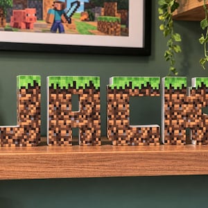 Könnte beinhalten: Holzbuchstaben, die "JACK" buchstabieren, mit einem pixeligen Design, das an das Spiel Minecraft erinnert. Die Oberseite jedes Buchstabens ist grün und simuliert Gras. Ein gerahmtes Bild zum Thema Minecraft und ein grünes Creeper-Plüschtier befinden sich im Hintergrund.