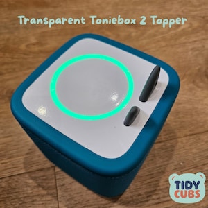 Könnte beinhalten: Ein quadratischer, türkis-weißer Toniebox 2 Topper mit einem leuchtend grünen Kreis und zwei grauen Knöpfen. Der Text "Transparent Toniebox 2 Topper" steht oben. Das Tidy Cubs-Logo befindet sich unten rechts.
