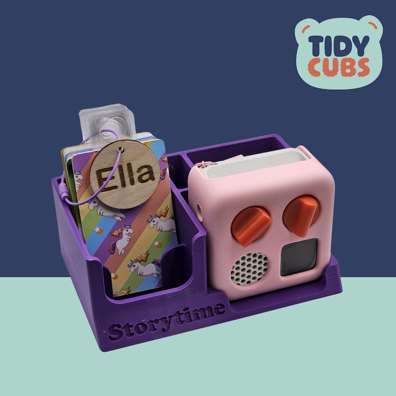 May include: Purple plastic organizer with the word "Storytime" on the front. The organizer has two compartments, one for a pink toy with orange buttons and a speaker, and the other for cards with a unicorn design. A wooden tag with the name "Ella" is attached to the cards.