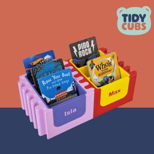 Puede incluir: Un organizador de almacenamiento infantil multicolor con el texto "TIDY CUBS". El organizador tiene compartimentos etiquetados "Isla" y "Max", que contienen libros y tarjetas. Los libros incluyen "Row Your Boat" y "Who's in Your Audio Book?"