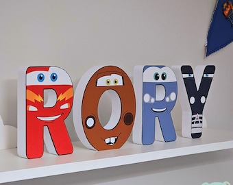 May include: Four white-edged wooden letters spelling "RORY" are displayed on a shelf. Each letter is designed to resemble a cartoon car, with vibrant colors including red, brown, blue, and black. The letters are on a white shelf.