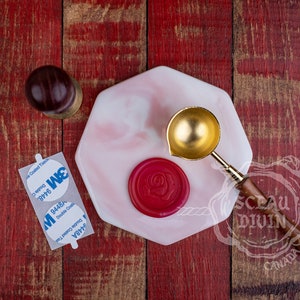 May include: A gold wax seal spoon with a wooden handle, a red wax seal with a rose design, and a white marble-like coaster on a red wooden surface. The coaster has the text "SCAU DIVIN CANADA" on it.