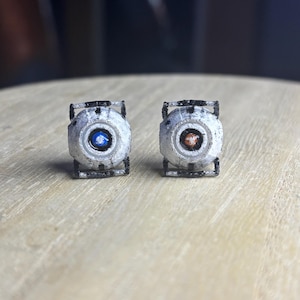 May include: A pair of small, spherical stud earrings featuring a textured white finish with black frame details. One earring has a central blue circular accent, while the other earring features a central orange circular accent, both with black borders.