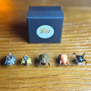 May include: A collection of five miniature sculptures, each with unique designs and colors, are displayed on a wooden surface. A dark blue box with a white logo is in the background. The sculptures include a metallic blue insect, a yellow and black insect, a brown insect, a red and orange insect, and a black insect.