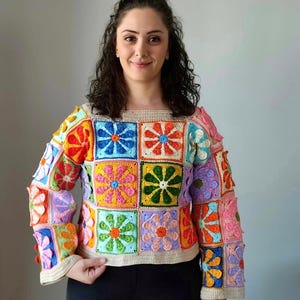 May include: A colorful crocheted sweater with a square pattern of flowers in various colors, including pink, orange, green, blue, and yellow.