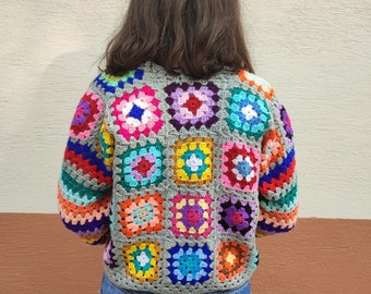 Puede incluir: Un cárdigan de crochet colorido con un patrón de cuadrados de la abuela en tonos de azul, verde, rojo, amarillo, rosa y morado. El cárdigan está hecho con un hilo gris y tiene un ajuste holgado.