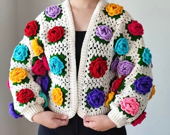 Puede incluir: Una chaqueta de punto de ganchillo blanca con un diseño floral colorido. La chaqueta presenta una variedad de colores, incluyendo rojo, rosa, azul, amarillo y verde. Las flores están tejidas en crochet en 3D y están unidas a la chaqueta.