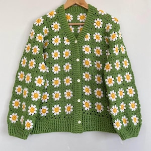 Könnte beinhalten: Grüne Häkeljacke mit einem Gänseblümchenmuster und Knöpfen.