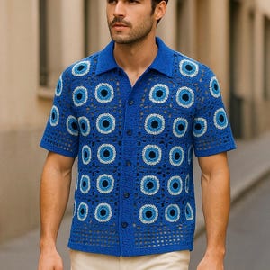 Puede incluir: Una camisa azul de crochet con un patrón de ojo blanco y azul. La camisa tiene un frente abotonado y mangas cortas.